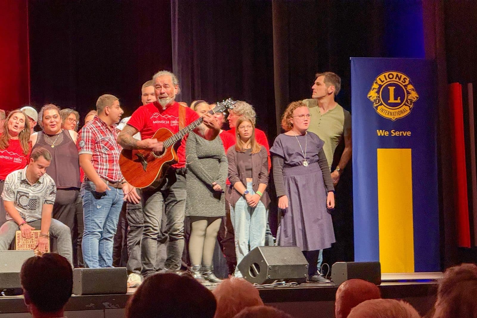 Der Musiker Alfons Hasenknopf mit einer Gitarre, im Hintergrund der Chor mit behinderten Menschen.